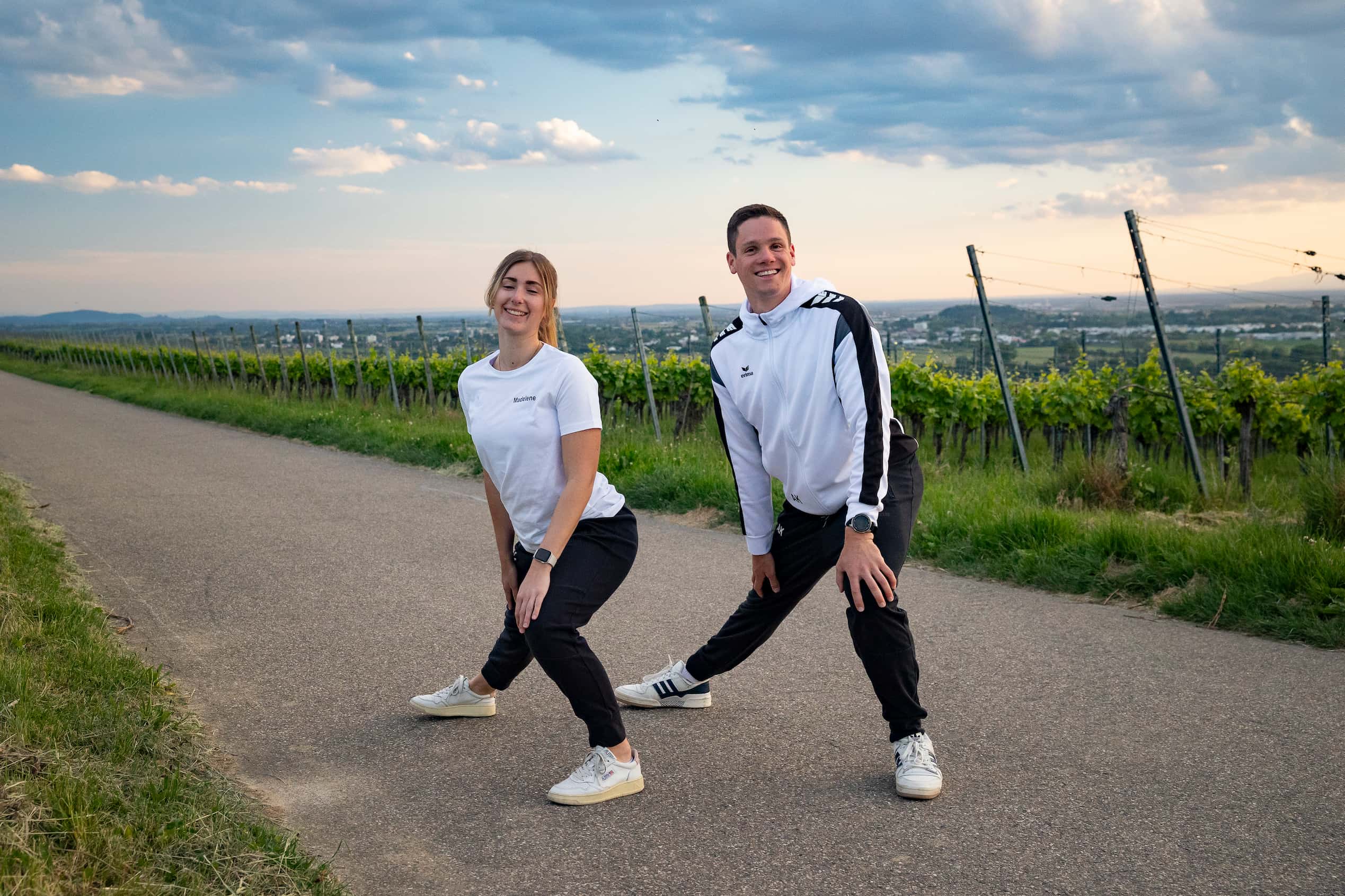 kontakt Zwei Vereinsmitglieder in weißen TV-Shirts beim Dehnen auf einem Weg durch die Weinberge